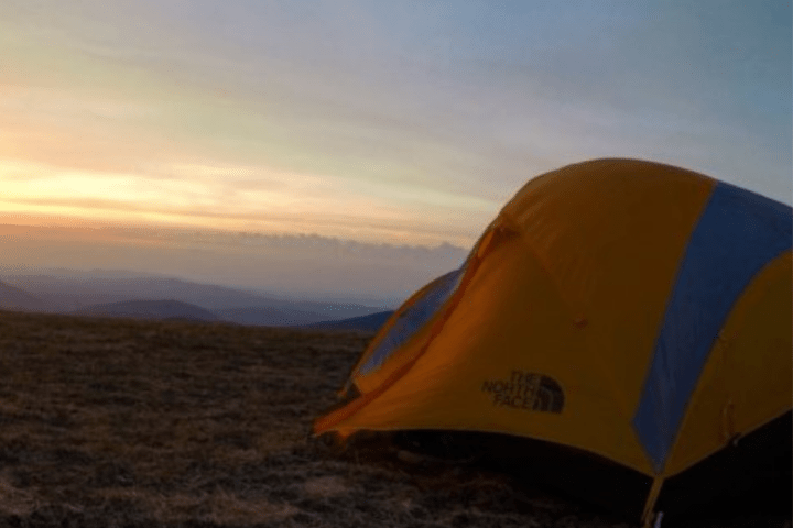 Orange tent on grassy field with mountains and sunrise in background.