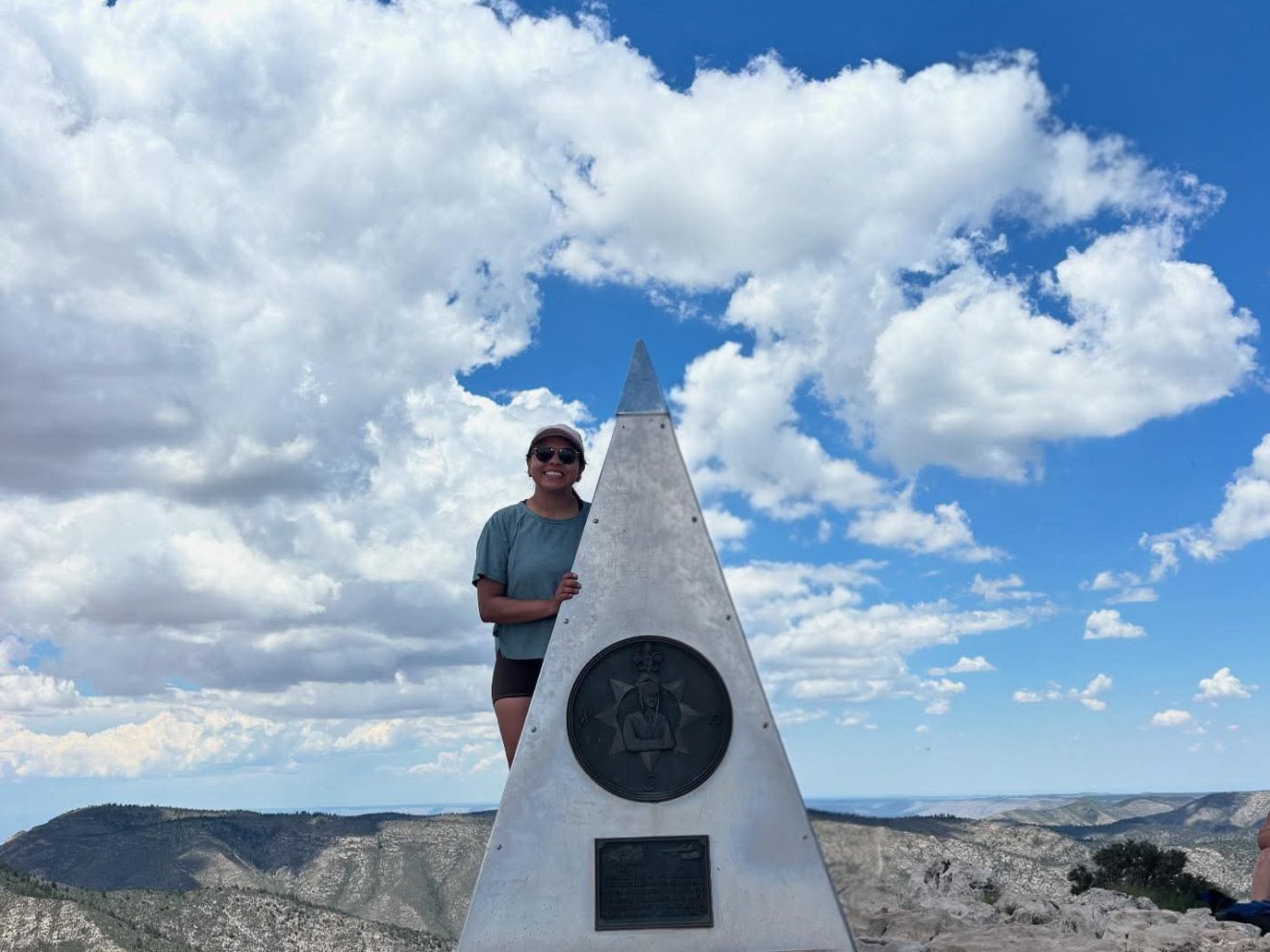 Person standing by triangular monument on rocky mountaintop under a blue sky with clouds.
