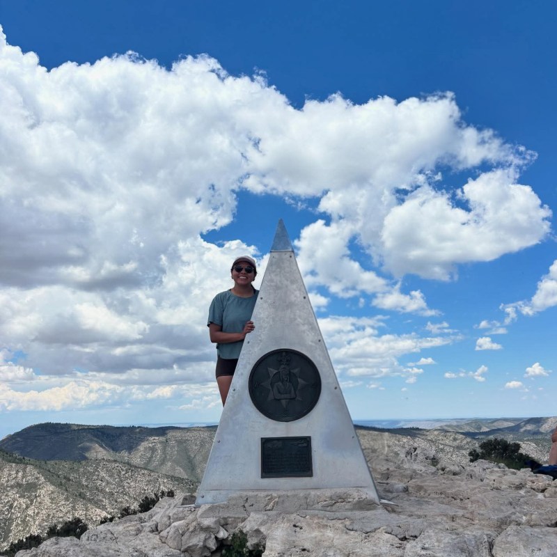 Person standing by triangular monument on rocky mountaintop under a blue sky with clouds.