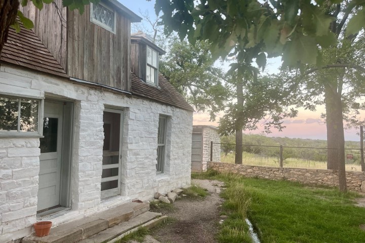 A rustic white stone house with wood accents, surrounded by trees and a grassy yard.