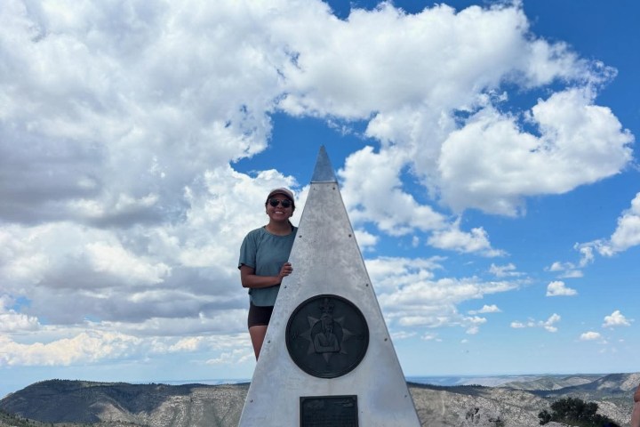 Person beside a triangular monument on a rocky mountain peak under a blue sky with scattered clouds.
