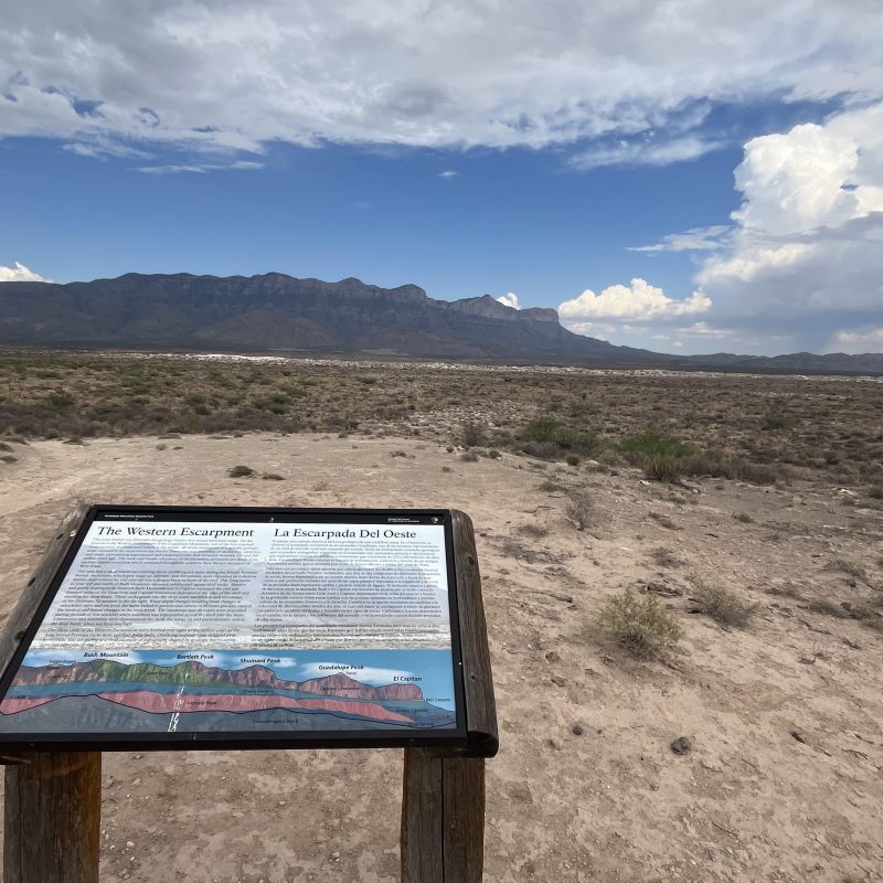 Information sign with mountain view under a cloudy blue sky in a desert landscape.