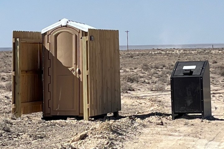 Portable toilet with wooden enclosure on a dirt road in a barren landscape.
