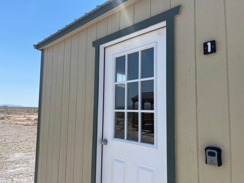 Small beige shed with a white door, keypad entry, and number 1, in a desert area.