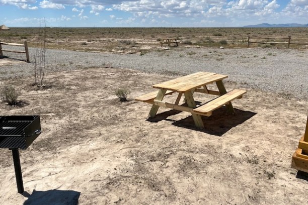 Wooden picnic table and grill in a desert area under a cloudy sky.