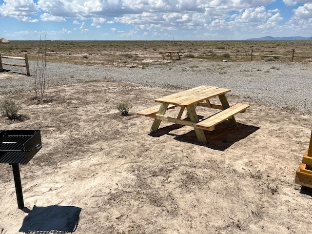 Wooden picnic table and grill in a desert area under a cloudy sky.