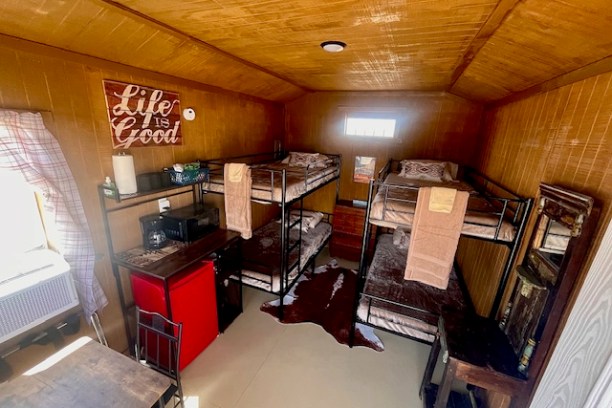 Interior with bunk beds, red fridge, microwave, table, and 'Life is Good' sign on wood-paneled walls.