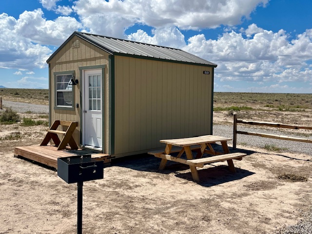 Small cabin with picnic table and grill in a desert landscape under a blue sky with clouds.