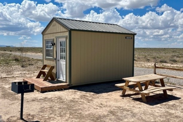 Small cabin with a metal roof, picnic table, and grill in a dry landscape under a cloudy sky.