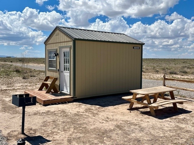 Small cabin with a metal roof, picnic table, and grill in a dry landscape under a cloudy sky.