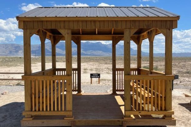 Wooden gazebo with a metal roof in a desert landscape, mountains in the background.