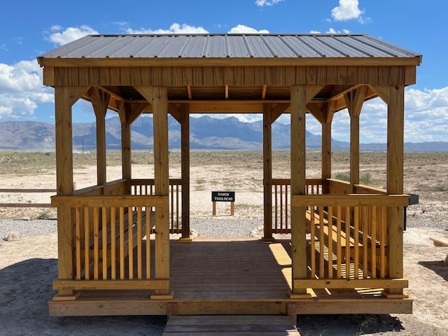 Wooden gazebo with a metal roof in a desert landscape, mountains in the background.