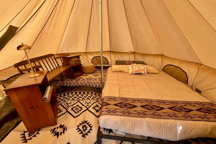 Interior of a cozy tent with a bed, wooden desk, lamp, and patterned rug.