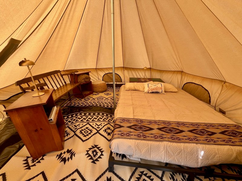 Interior of a cozy tent with a bed, wooden desk, lamp, and patterned rug.