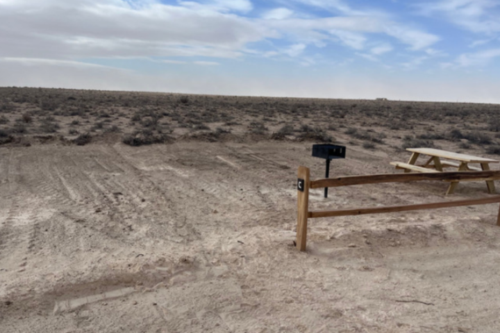 Desert landscape with a wooden fence, picnic table, and grill under a cloudy sky.