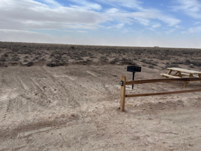 Desert landscape with a wooden fence, picnic table, and grill under a cloudy sky.