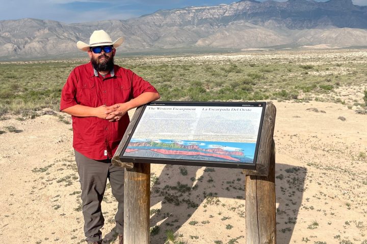 Man in red shirt and hat stands by informational sign in desert landscape with mountains.