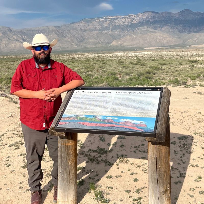 Man in red shirt and hat stands by informational sign in desert landscape with mountains.
