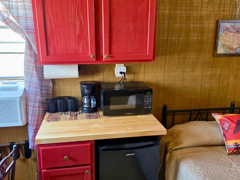 Small kitchen area with red cabinets, microwave, coffee maker, mini fridge, and a bed nearby.