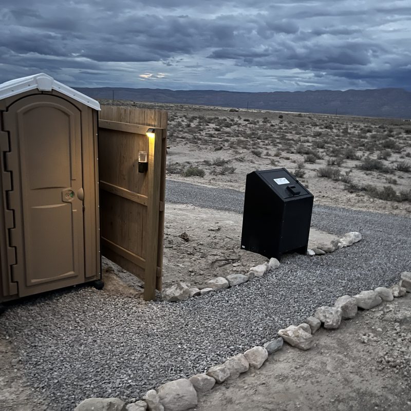 Portable toilet with light and trash bin on gravel path in desert landscape, under cloudy sky.