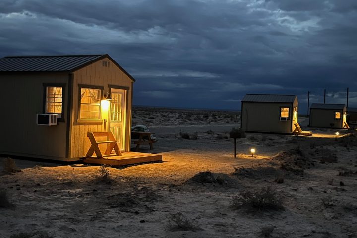 Small cabins with warm lights in a desert landscape under a cloudy sky at dusk.