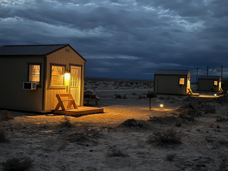 Small cabins with warm lights in a desert landscape under a cloudy sky at dusk.