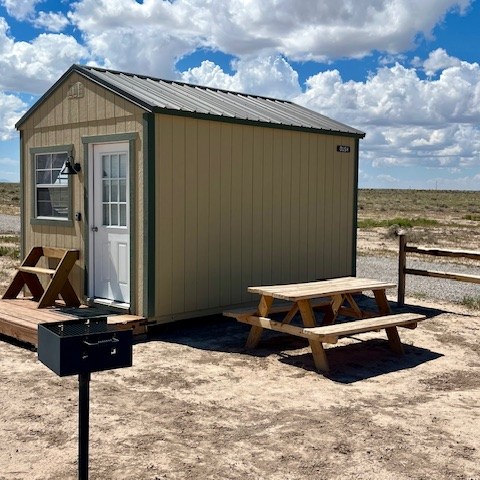 Small cabin with a picnic table and grill on a sandy lot under a blue sky with clouds.