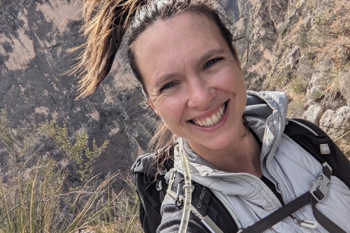 Smiling person with a backpack hiking on a mountainous trail with rocks and trees.