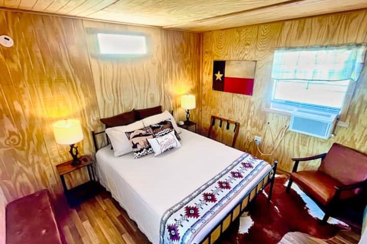 Cozy bedroom with wood paneling, queen bed, Texas flag, lamps, chairs, and sunlight from a window.