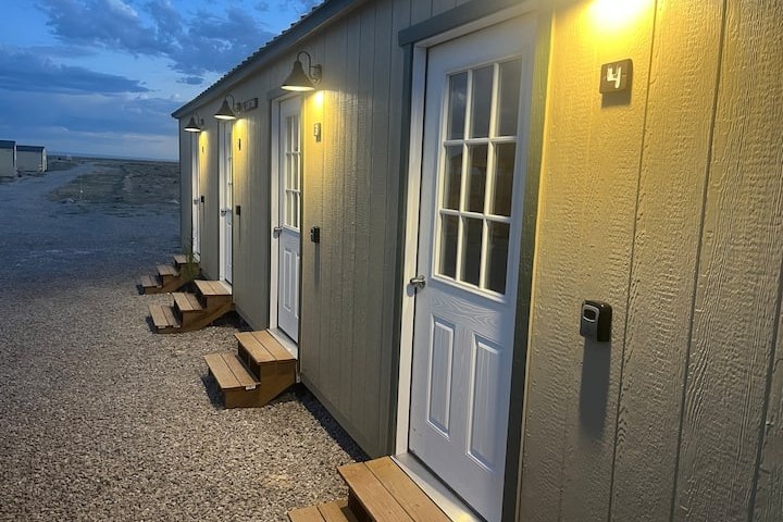 Small cabins with white doors and porch lights in evening light on a gravel path.