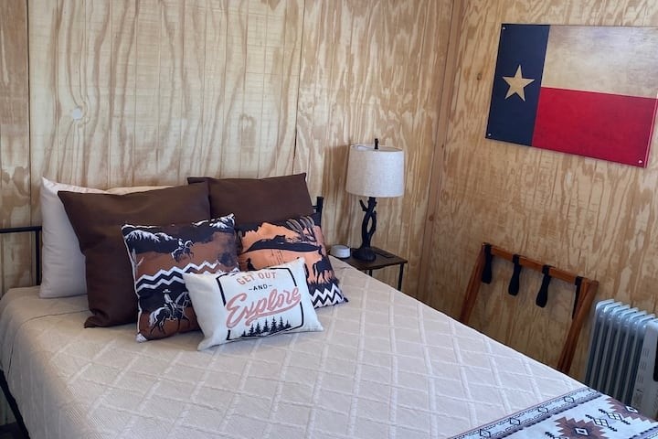 Cozy bedroom with wooden walls, a bed with decorative pillows, a small lamp, and a Texas flag on the wall.