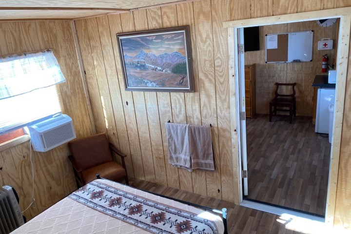 Wood-paneled room with bed, chair, window AC, painting, towel, and view into another room.