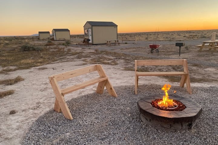 Two wooden benches by a fire pit with small cabins in the background at sunset.