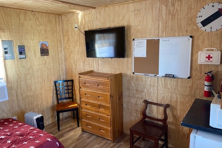 Small room with wooden walls, dresser, TV, chairs, whiteboard, wall clock, and emergency kit.